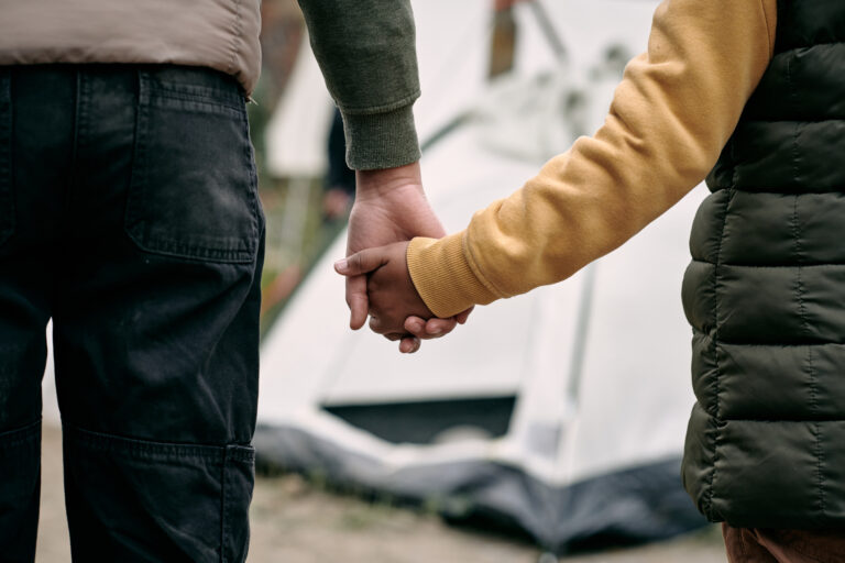 Rear view of unrecognizable parent of elder brother holding hand of child against tent in migrant camp