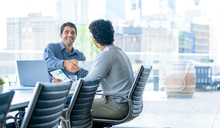 Two businessmen working on a digital tablet and laptop computer in the office in a board room or meeting room. They are shaking hands closing the deal and a large window behind them