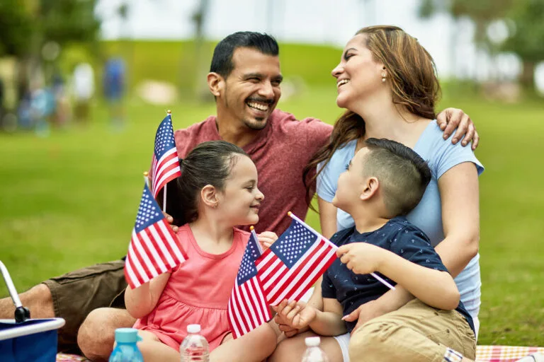 Smiling Hispanic American family with pre-adolescent children holding American flags while celebrating holiday at Miami park with picnic.